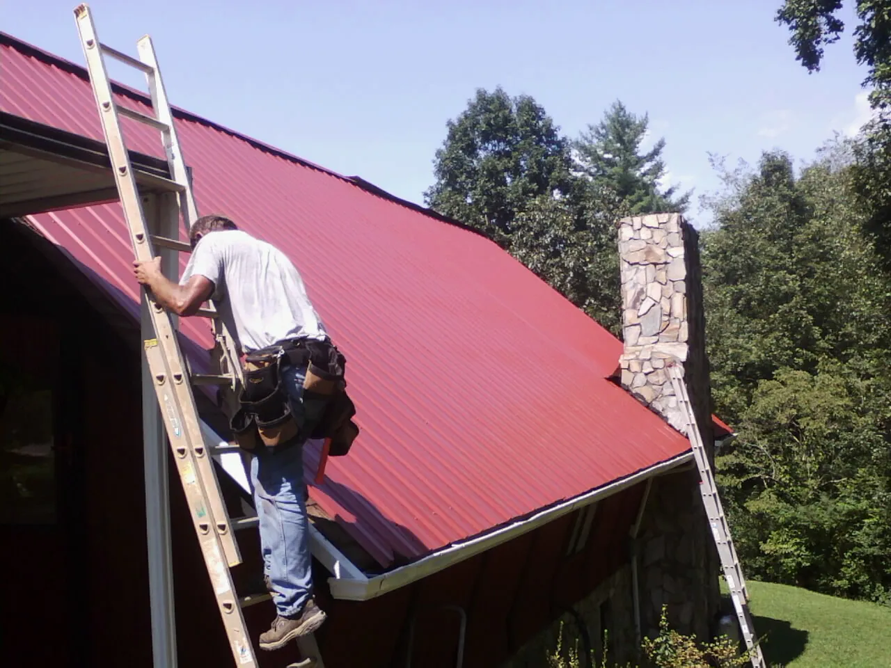 Roofer on a ladder beside a red metal roof and stone chimney — Soffit & Fascia Repair by Roofing Brothers in Forest Park